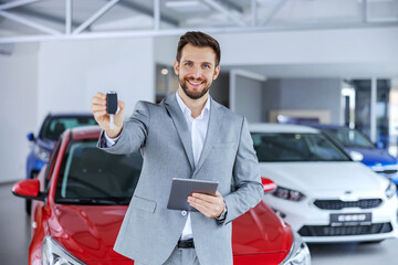 Car seller standing in car salon and showing keys on a new brand car which are ready to be sold.