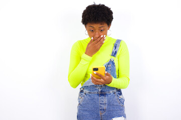 young African American woman with short hair wearing denim overall against white wall being deeply surprised, stares at smartphone display, reads shocking news on website, Omg, its horrible!