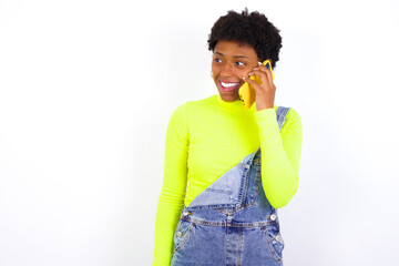Pleasant looking happy young African American woman with short hair wearing denim overal has nice telephone conversation and looks aside, has nice mood and smiles positively while talks via cell phone