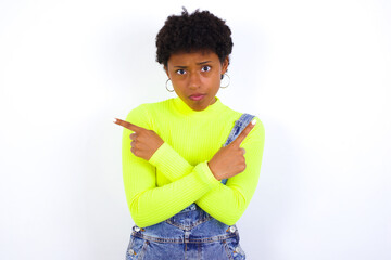 Serious African American female with curly bushy wears jeans overalls over white wall crosses hands and points at different sides hesitates between two items. Hard decision concept