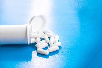 White cylindrical capsules on a matte blue background fall out of white tube. View from above. Medical concept of treatment, prevention and side effects.