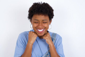 young African American woman with short hair wearing denim overall against white wall grins joyfully, imagines something pleasant, copy space. Pleasant emotions concept.