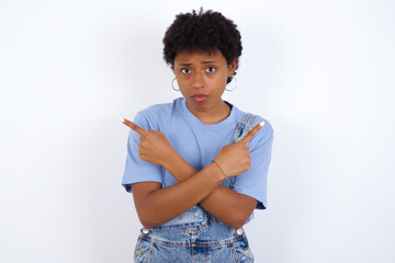 Serious young African American woman with short hair wearing denim overall against white wall crosses hands and points at different sides hesitates between two items. Hard decision concept