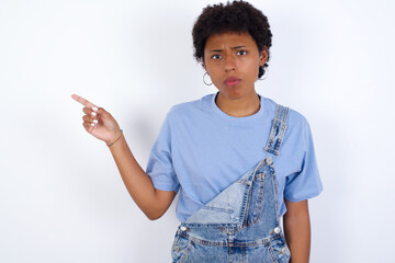 Serious young African American woman with short hair wearing denim overall against white smirks face points away on copy space shows something unpleasant. Look at this advertisement. Big price concept