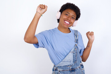 Carefree young African American woman with short hair wearing denim overall and toothy smile raises arms dances carefree moves with rhythm of music listens music from playlist via headphones