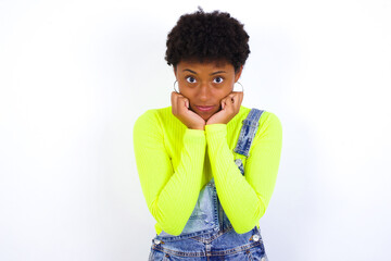 Portrait of sad African American female with curly bushy wears jeans overalls over white wall hands face