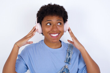 Joyful young African American woman with short hair wearing denim overall against white wall sings song keeps hand near mouth as if microphone listens favorite playlist via headphones