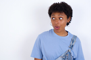 Shocked young African American woman with short hair wearing denim overall against white wall look empty space with open mouth screaming: Oh My God! I can't believe this.