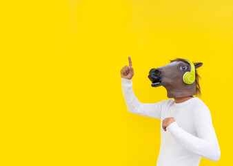 Man with horse head mask and headphones using smartphone ,points with finger up on blank wall ,indicates copy space isolated on yellow background.
