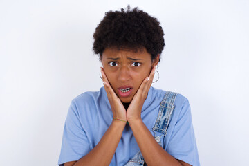 Fototapeta na wymiar Upset young African American woman with short hair wearing denim overall against white wall touching face with two hands