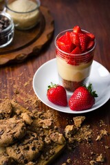 Strawberry trifle dessert with pudding and cookies on dark wooden background with glass jars