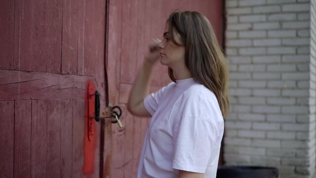 Side view of young beautiful Caucasian brunette woman knocking in big red door outdoors. Portrait of serious charming lady in industrial suburb on summer day