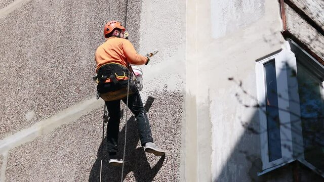 Dirty industrial climber in orange clothes working near buildings outside. Worker hanging on ropes and painting the wall with sealant