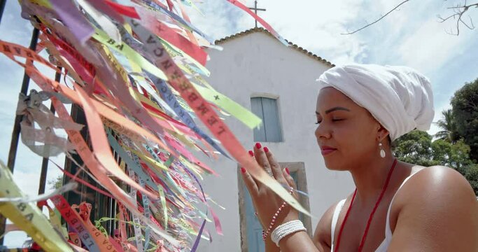 Brazilian woman dressed in the traditional Bahian costume of the Umbanda religion saying a prayer. Colored ribbons of religious promises in devotion to Senhor do Bonfim in Bahia, Brazil. 6K.