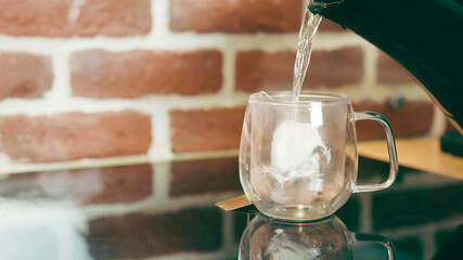 Making tea in a bag in a transparent glass cup on the kitchen table on a background of red brick wall. Tea in a bag is filled with hot water from the kettle.