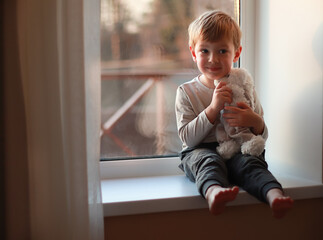 A little smiling boy sits on a windowsill in the evening light and hugs a teddy bear. International Children's Day