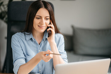 Young pretty woman talking on video call and waving hand while sitting at table in the homeoffice room