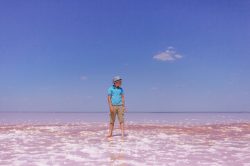 happy teenager in blue T-shirt, beige shorts and hat standing on an amazing pink lake with sea water and blue sky