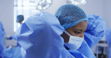 African american female surgeon wearing surgical cap, putting face mask on in operating theatre