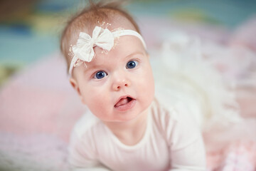 a cute baby looks into the camera with a banter on his head and in white clothes. The mouth is open and the tongue is visible. Blue eyes and fluffy hair