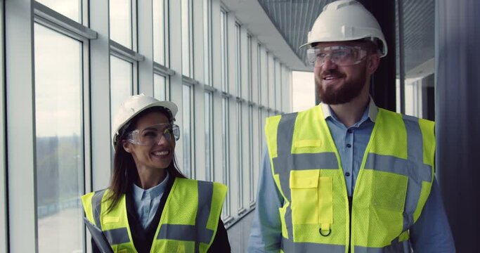 Smiling woman architect and chief male engineer walking at construction site of unfinished commercial center, having pleasant conversation, looking proud and satisfied.