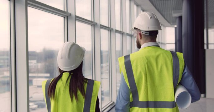 Back view of woman architect and chief male engineer walking at construction site of unfinished commercial center with panoramic windows, discussing work process.