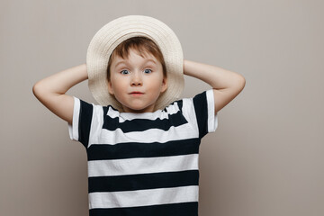 Studio portrait of a charming cute boy in a straw hat with a surprised face holding his hands behind his head on a gray background.