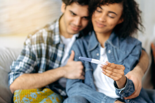 Joyful mixed race married couple waiting to be replenished. Husband and wife are sitting on the sofa, african american girl holding a positive pregnancy test in hand, they preparing to become - Powered by Adobe