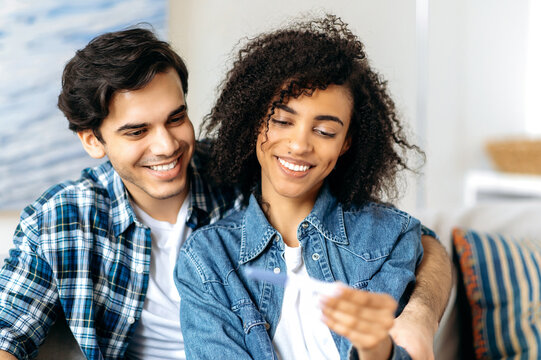 Young happy mixed race married couple sitting on sofa looking at positive pregnancy test enjoying good news, getting ready to become parents,african american girl holds positive pregnancy test in hand