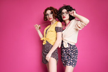 Obraz na płótnie Canvas Two cute girl friends woman looking like nerd accountants standing on a pink background. They wear curly brunette wigs and unstylish retro casual outfits.