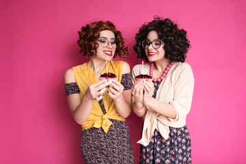 Two cute girl friends woman looking like nerd accountants standing on a pink background. They wear curly brunette wigs and unstylish retro casual outfits. Berry cupcake with candle in their hands.