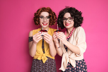 Two cute girl friends woman looking like nerd accountants standing on a pink background. They wear curly brunette wigs and unstylish retro casual outfits. Berry cupcake with candle in their hands.