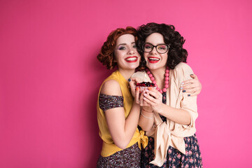 Two cute girl friends woman looking like nerd accountants standing on a pink background. They wear curly brunette wigs and unstylish retro casual outfits. Berry cupcake with candle in their hands.
