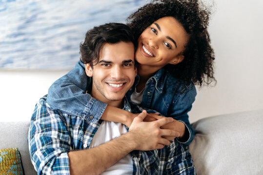 Portrait joyful happy mixed race couple, pretty african american curly girl tenderly hugging her hispanic boyfriend sitting on sofa, wearing casual clothes in living room, looking at camera, smiling