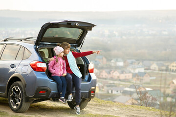 Side view of mother with daughter sitting in car trunk and looking on nature. Concept of resting with family on fresh air.