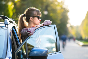 Young woman driver enjoying warm summer day standing beside her car on city street. Travelling and vacation concept.