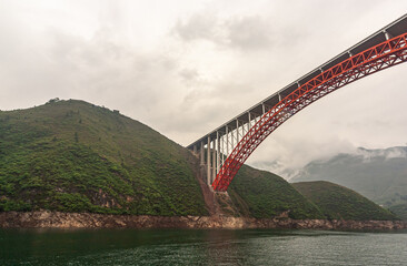 Obraz premium Wushan, China - May 7, 2010: Dragon Gate Gorge on Daning River. Red bow with flat white concrete road surface bridge over dark green water under cloudscape. Mountainous shorelines with green foliage.