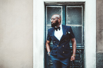 A handsome dapper bald black guy with a beard, in an elegant dark-blue suit with a bow-tie and glasses, has just left a house through a peeled antique door and looking aside - obrazy, fototapety, plakaty