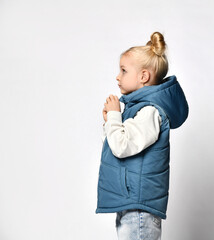 Side view of a little girl in warm outerwear blue sleeveless vest, jeans and a sweatshirt, posing for the camera isolated on white background. Children's fashion clothing and fashion advertising