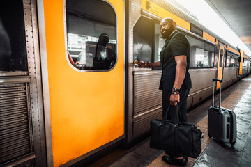 A wide-angle view of a mature bald bearded black man entrepreneur in a suit and with luggage bags, pushing the button to open a vivid yellow door of a high-speed train to start his business travel