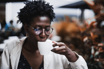 Portrait of a young glamorous black female in classy spectacles drinking a hot tea in a street cafe; ravishing African woman in glasses drinking a delicious latte from the cup in an outdoor restaurant - obrazy, fototapety, plakaty