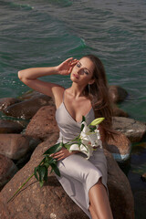 Elegant woman in grey silk dress sitting at stones at sea coast against azure water and holding...