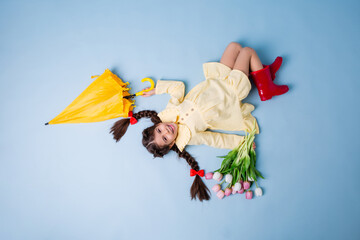 happy little girl in rubber boots and a summer dress holds a yellow umbrella and a bouquet of tulips, lies on a blue background in the studio, top view, place for text
