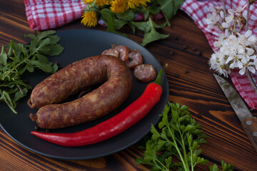 Smoked sausage ring with sliced pieces on black plate with red chili pepper and fresh herbs on...