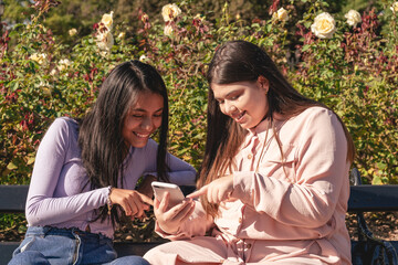Two latin female friends sitting on a bench looking at cell phones in a park