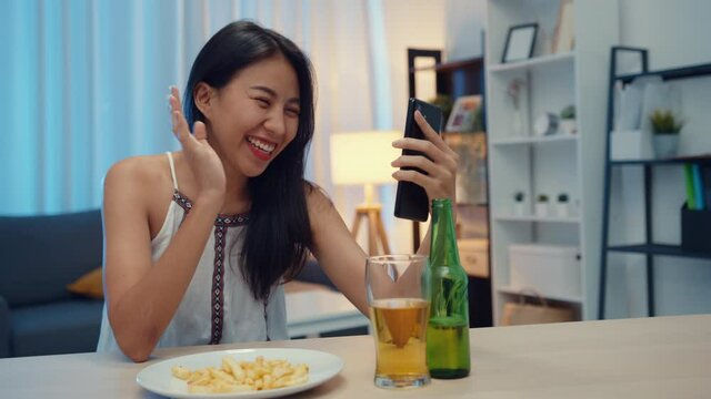 Young Asia lady drinking beer having fun happy moment night party New Year event online celebration via video call by phone at home at night. Social distancing, quarantine for coronavirus prevention.