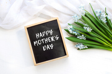 Fresh blue spring flowers Scilla siberica and wooden blackboard with sample text Happy Mothers Day white table.