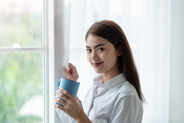 Asian woman drinking a cup of coffee and looking through the glass window after wake up in the morning