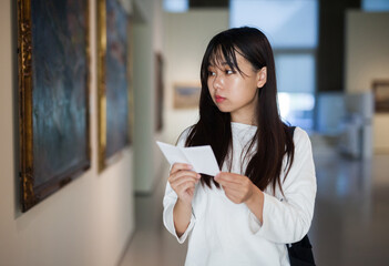 Fototapeta premium Chinese woman standing with guide-book in art museum near the painting in baguette