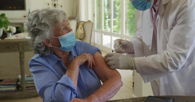 Caucasian senior male doctor wearing face mask injecting covid-19 vaccine into senior woman at home